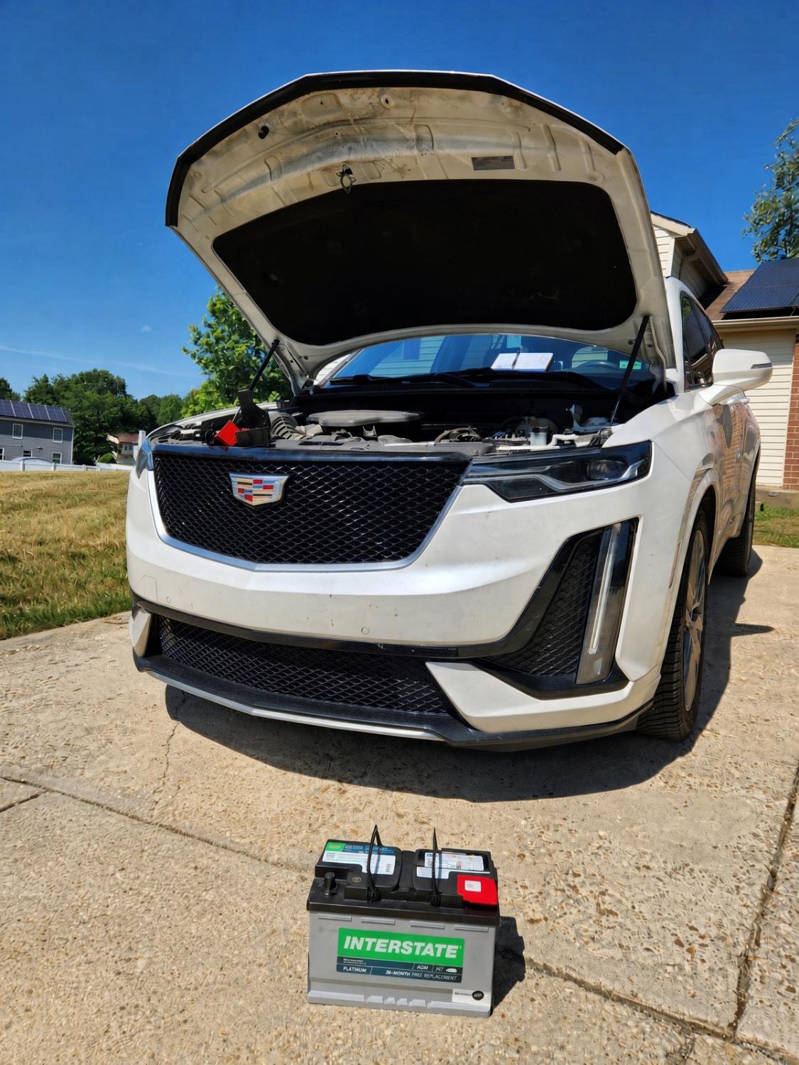 White Cadillac with the hood open and a new battery on the driveway.